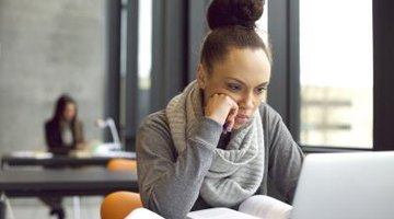A college student sits at a desk with open books, while looking at a laptop screen